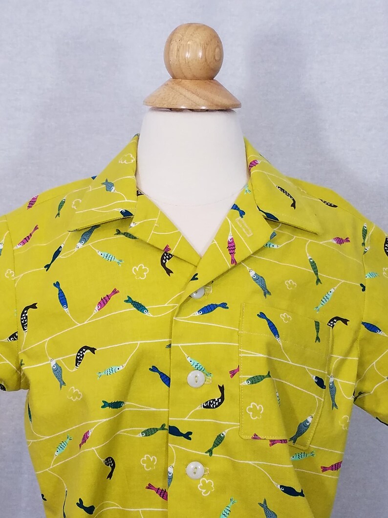 May include: A short-sleeved button-down shirt in a bright yellow-green color, adorned with a repeating pattern of colorful fish and white wavy lines. The shirt has a single chest pocket and white buttons.