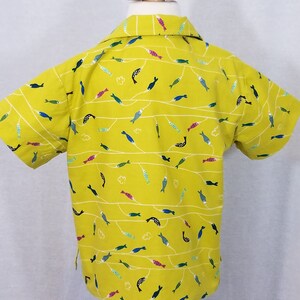 May include: A short-sleeved yellow shirt with a repeating pattern of colorful fish and white wavy lines. The shirt is displayed on a mannequin and has a classic collar.