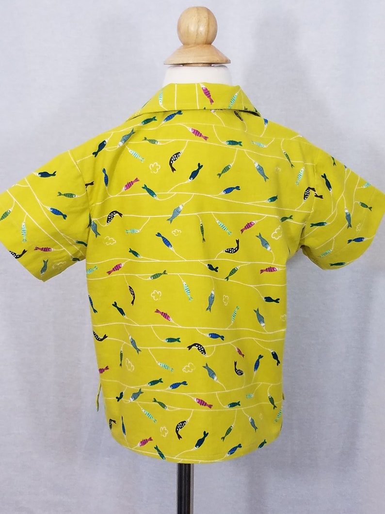 May include: A short-sleeved yellow shirt with a repeating pattern of colorful fish and white wavy lines. The shirt is displayed on a mannequin and has a classic collar.
