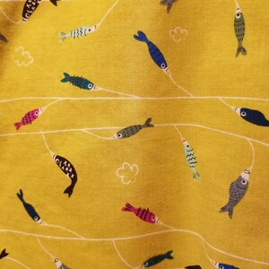 May include: Close-up of a yellow fabric with a repeating pattern of colorful fish and white lines. The fish are in various colors, including blue, pink, green, and black. The design includes small white cloud shapes.