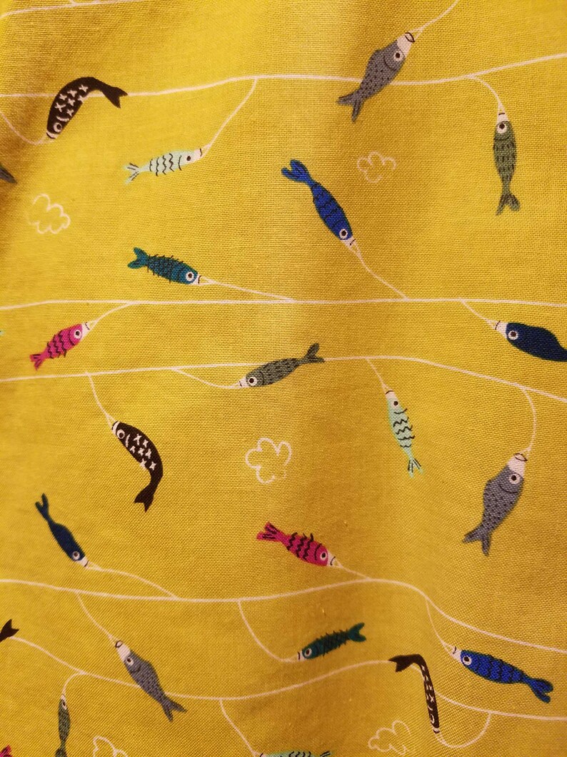May include: Close-up of a yellow fabric with a repeating pattern of colorful fish and white lines. The fish are in various colors, including blue, pink, green, and black. The design includes small white cloud shapes.