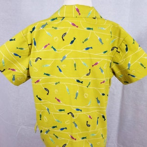 May include: A short-sleeved, yellow button-up shirt with a repeating pattern of colorful fish. The fish are in various colors, including blue, pink, and green, and are arranged along white, wavy lines. The shirt is displayed on a mannequin.