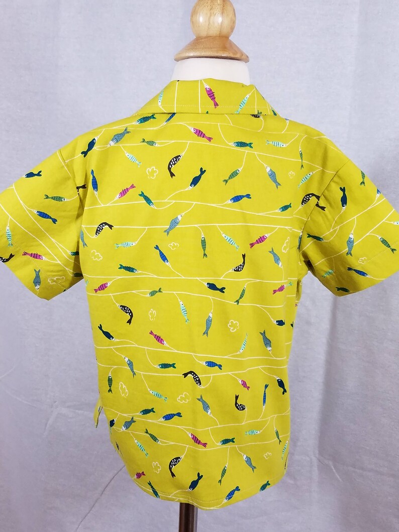 May include: A short-sleeved, yellow button-up shirt with a repeating pattern of colorful fish. The fish are in various colors, including blue, pink, and green, and are arranged along white, wavy lines. The shirt is displayed on a mannequin.
