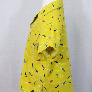 May include: A yellow button-down shirt with a playful fish pattern. The shirt features short sleeves, a collar, and a pocket. The fish are in various colors, including blue, green, and pink, and are arranged on white wavy lines.