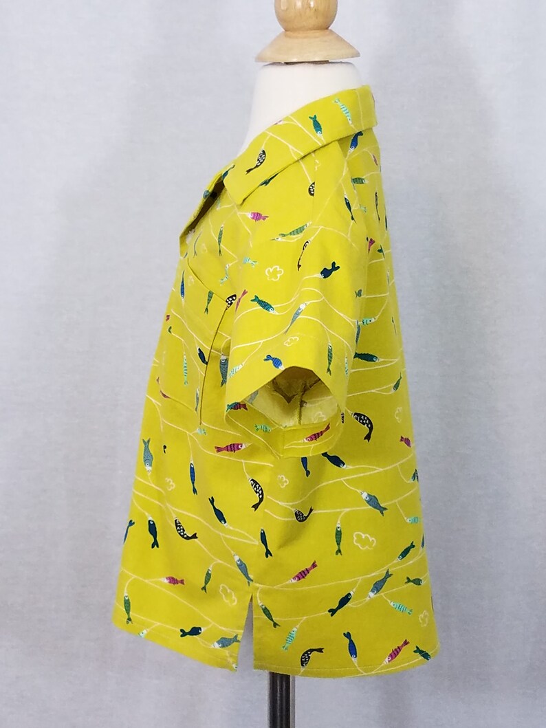 May include: A yellow button-down shirt with a playful fish pattern. The shirt features short sleeves, a collar, and a pocket. The fish are in various colors, including blue, green, and pink, and are arranged on white wavy lines.