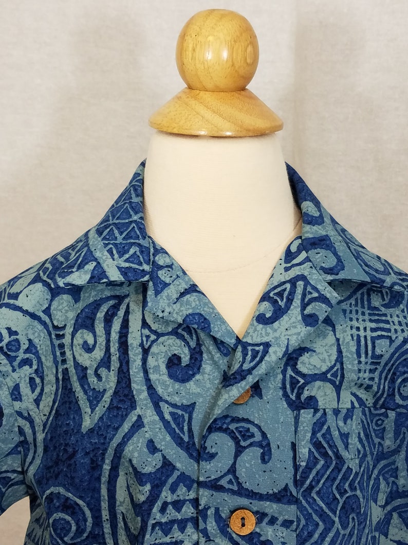 Puede incluir: Una camisa de manga corta con estampado azul y verde con cierre de botones de madera. La camisa tiene un dise&ntilde;o tradicional hawaiano.