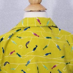May include: A yellow collared shirt with a repeating pattern of colorful fish. The fish are in various colors, including blue, pink, and green, and are connected by white lines. The shirt is displayed on a wooden mannequin head.