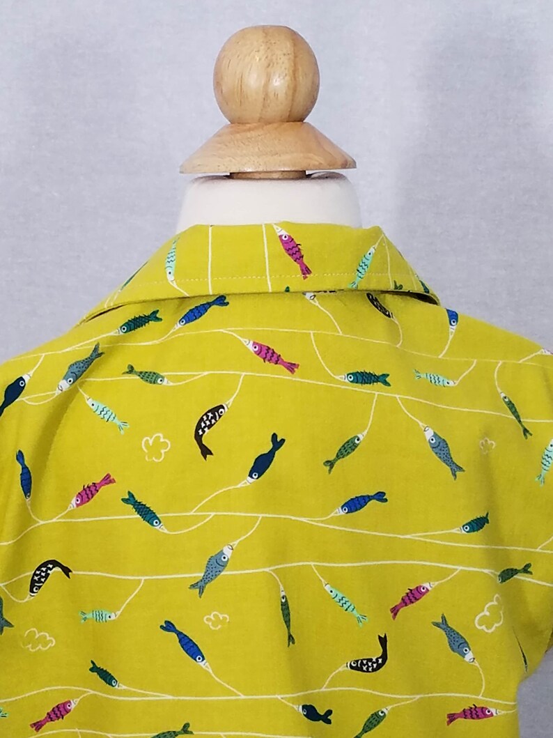 May include: A yellow collared shirt with a repeating pattern of colorful fish. The fish are in various colors, including blue, pink, and green, and are connected by white lines. The shirt is displayed on a wooden mannequin head.