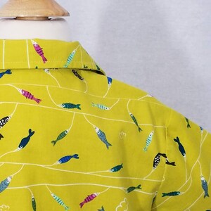 May include: A yellow button-down shirt with a repeating pattern of colorful fish and white lines. The fish are in various colors, including pink, blue, green, and black. The shirt is on a wooden mannequin head.