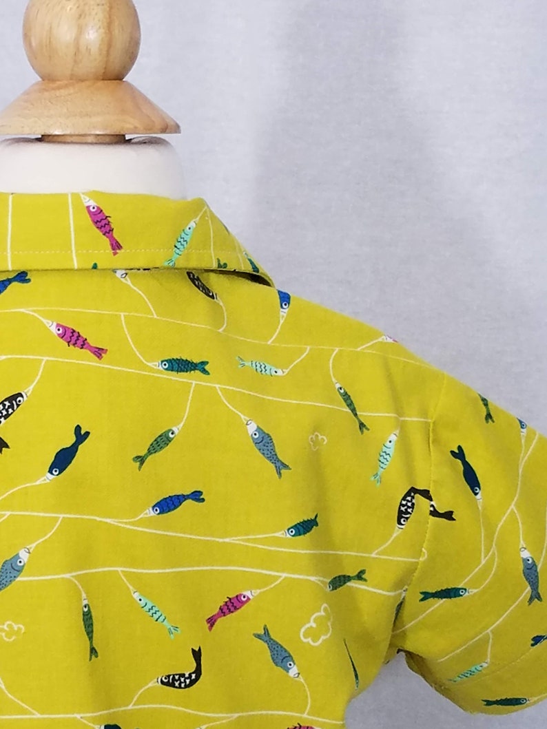 May include: A yellow button-down shirt with a repeating pattern of colorful fish and white lines. The fish are in various colors, including pink, blue, green, and black. The shirt is on a wooden mannequin head.