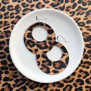 May include: A pair of large, round, hoop earrings with a leopard print design. The earrings are made of leather and have a silver hook fastening.