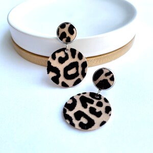 May include: A pair of dangle earrings featuring a leopard print design. The earrings are made of metal and have a round shape. The top part of the earrings is a smaller circle, and the bottom part is a larger circle.