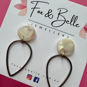 May include: A pair of Fox & Belle Jewellery earrings. Each earring has a pearlescent, circular stud attached to a teardrop-shaped, silver-toned frame. The brand name and social media icons are visible on the white card.