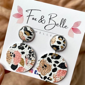May include: A pair of dangle earrings featuring a white, black and gold leopard print design. The earrings have a smaller round top and a larger round bottom. The earrings are displayed on a white card with the text "Fox & Belle Jewellery".