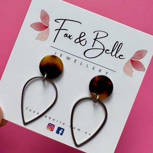May include: Earrings featuring a tortoiseshell design and teardrop-shaped frames. The earrings are displayed on a white card with the text "Fox & Belle Jewellery" and social media icons. The background is pink.