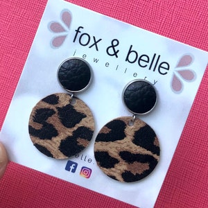 May include: A pair of dangle earrings featuring a black leather circle and a larger circle with a leopard print pattern. The earrings are displayed on a white card with the text "fox & belle jewellery" and social media icons.