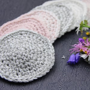 Puede incluir: Un juego de posavasos de ganchillo en tonos rosa y gris. Los posavasos están dispuestos en semicírculo con un ramo de flores silvestres en primer plano.