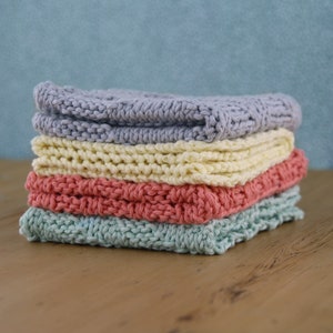 May include: A stack of four knitted dishcloths in different colors: light blue, cream, coral, and light green. The dishcloths are folded and stacked on a wooden surface.