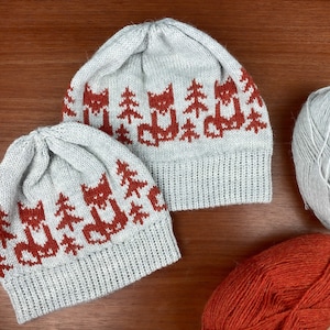 May include: Two grey knitted hats with a red fox and pine tree pattern. The hats are on a wooden surface with two balls of yarn, one grey and one orange.