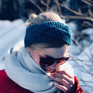 May include: A woman wearing a teal blue knitted headband and a white scarf. She is wearing sunglasses and a red jacket.