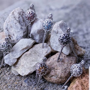 May include: Five silver key-shaped necklaces with a round, ornate, floral design. Each necklace has a different colored stone inside the floral design. The necklaces are displayed on a bed of gray rocks.