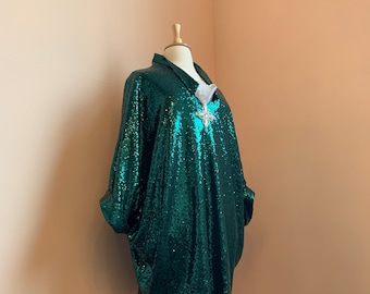 Puede incluir: Una blusa verde de lentejuelas con un broche blanco adornado. La blusa tiene un corte holgado y un cuello.