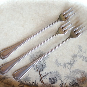 May include: Three silver-toned metal cocktail forks with long, slender handles and three tines. Each fork features a simple, smooth design with a slightly flared, rounded end on the handle, providing a clean and functional appearance for dining.