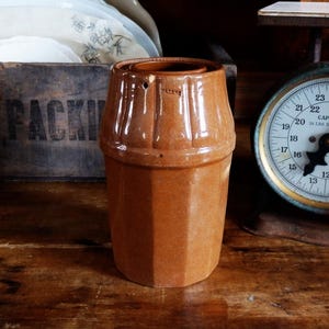 May include: A vintage brown ceramic container with a lid, featuring a hexagonal base and a ridged top. The container is set on a wooden surface, with a scale in the background. The container is likely for kitchen use.