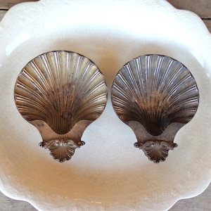 May include: Two antique, shell-shaped dishes with a metallic finish. The dishes have a fan-like design and are set on a white, scalloped-edge plate. The dishes have a decorative, ornate base.