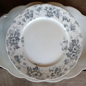 May include: Two white ceramic plates. The top plate has a floral design in gray and white. The bottom plate is larger, with a scalloped edge and a subtle embossed pattern. The plates are on a wooden surface.