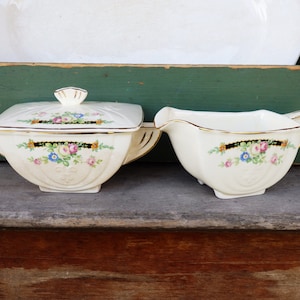 May include: A vintage white porcelain sugar and creamer set with gold trim and floral designs. The creamer has a lid and both pieces have a square shape.