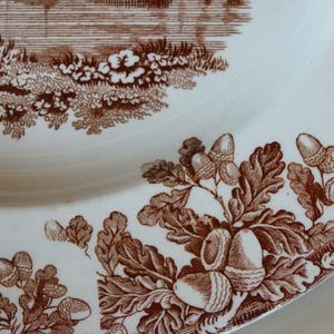 Może przedstawiać: Biały ceramiczny talerz z brązowym wzorem transferowym. Wz&oacute;r przedstawia żołędzie, liście dębu i krajobraz. Talerz ma lekko zakrzywioną krawędź i błyszczące wykończenie.