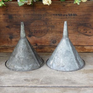 May include: Two conical metal funnels with a weathered, galvanized finish. Each funnel features a wide circular opening at the top that tapers down into a narrow cylindrical spout. The metal surfaces show variations in texture and gray tones.