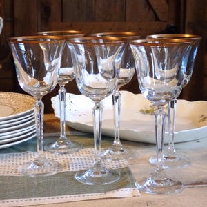 May include: A collection of six clear wine glasses with gold rims, arranged on a table with a stack of plates and a decorative serving dish. The glasses have long stems and are set on a green and white striped tablecloth.