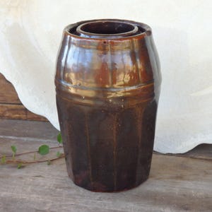 May include: A cylindrical ceramic crock featuring a dark brown, high-gloss glaze and a vertical ribbed texture along the exterior body. The vessel has a wide, open top with a thick, rounded rim and a slightly tapered shape toward the base.