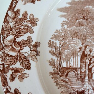 Może przedstawiać: Biały ceramiczny talerz z brązowym wzorem obramowania z żołędzi i liści dębu. Talerz przedstawia szczeg&oacute;łową brązową scenę krajobrazową z drzewami, mostem i elementami architektonicznymi.