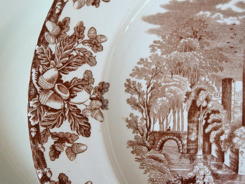 Może przedstawiać: Biały ceramiczny talerz z brązowym wzorem obramowania z żołędzi i liści dębu. Talerz przedstawia szczeg&oacute;łową brązową scenę krajobrazową z drzewami, mostem i elementami architektonicznymi.