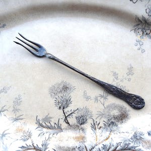 May include: A vintage silver fork with three tines, resting on a floral patterned plate. The fork has an ornate handle with floral detailing. The plate features a cream background with gray and blue floral designs, creating a classic aesthetic.
