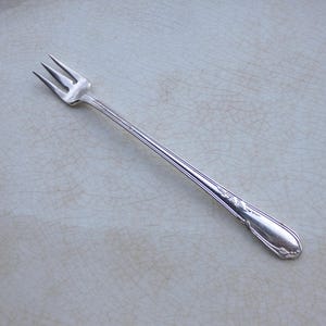May include: A silver-toned metal cocktail fork featuring three tines and a slender, elongated handle. The handle is decorated with a subtle, embossed floral motif near the base, showcasing a polished, reflective finish throughout the entire piece.