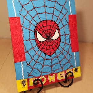 Può includere: Un arazzo da parete trapuntato rosso, blu e giallo con un disegno a ragnatela. Al centro della ragnatela è presente una maschera di Spiderman ricamata in rosso e nero. Le parole "With Great Power" sono ricamate nella parte superiore e inferiore della ragnatela.