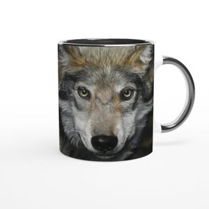 Könnte beinhalten: Eine Keramik-Kaffeetasse mit einem detaillierten Porträt eines Wolfsgesichts. Der Wolf hat graues und braunes Fell, grüne Augen und eine schwarze Nase. Die Tasse hat einen silbernen Rand und Henkel. Der Hintergrund ist weiß.
