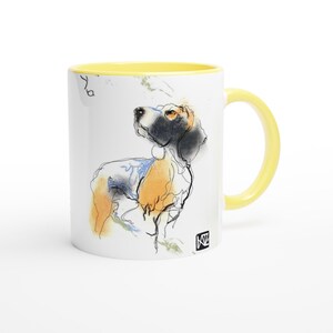 Könnte beinhalten: Weiße Keramiktasse mit gelbem Innenraum und Henkel. Die Tasse zeigt eine Aquarellillustration eines Hundes in Schwarz, Orange und Blau. Der Hund blickt nach oben. Eine kleine Künstlermarke ist sichtbar.