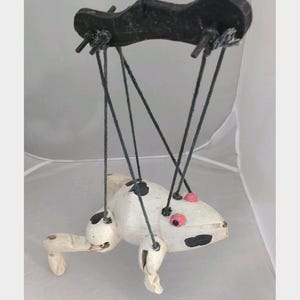 May include: A handmade wooden puppet with a white body, black spots, and pink eyes. The puppet is suspended by black strings attached to a black control bar. The puppet's limbs are articulated, and it appears to be a frog or similar creature.