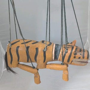 May include: A wooden tiger puppet with black stripes on a tan body, suspended by strings. The puppet has articulated limbs and a black tail. The head features painted details, including a black collar.
