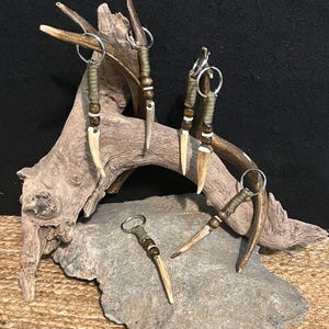May include: Five antler keychains with brown and white beads and a brown cord. The keychains are displayed on a piece of driftwood.
