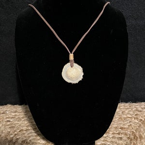 May include: A brown leather cord necklace with a round, white, and brown pendant. The pendant is made of a natural material, possibly bone or shell.