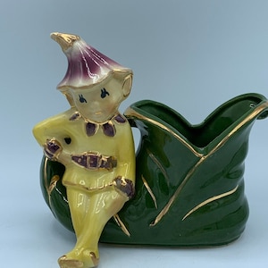 May include: A ceramic figurine of a yellow elf with a purple hat sitting on a green leaf shaped planter. The planter has gold accents.