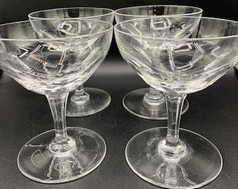 May include: Four clear champagne coupe glasses with etched designs. The glasses have a wide, shallow bowl, a slender stem, and a round base. The glasses are arranged in a square formation against a dark background.