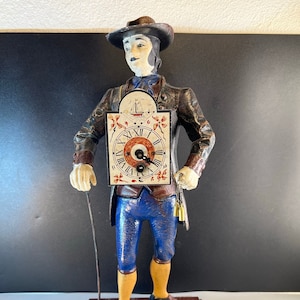 May include: A vintage clock figurine of a man in a blue suit and brown hat holding a clock face with Roman numerals. The clock face is surrounded by a decorative frame with a sailboat and flowers. The figurine is standing on a black and brown base.