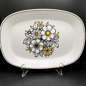 May include: White oval serving platter with a black trim and a floral design. The flowers are white, yellow, and gray with black outlines. The platter is displayed on a clear stand.